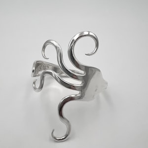 May include: A silver-colored fork-shaped bracelet with curved tines and a unique, artistic design. The bracelet is made of metal and has a polished finish. The tines are bent into decorative swirls.