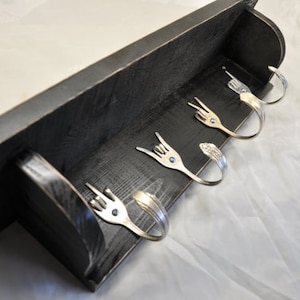 May include: A rustic, black wooden shelf with four decorative metal fork hooks. The forks are bent into various hand gestures. The shelf is designed for wall mounting and adds a unique touch to any space.