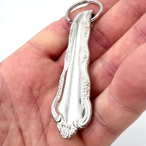 Könnte beinhalten: Schlüsselanhänger im Besteck-Design. Der Schlüsselanhänger hat ein löffelförmiges Design mit floralen und perlenartigen Details. Der Löffel ist an einem silbernen Metallring befestigt. Der Löffel besteht aus einem glänzenden, reflektierenden Metall.