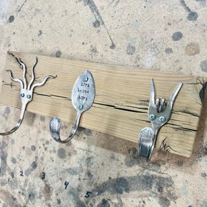 Peut inclure: Un porte-manteau mural en bois avec trois crochets fabriqués à partir de couverts réutilisés. Les crochets sont en forme de pieuvre, de cuillère avec les mots "Live Laugh Love" gravés dessus et de fourchette en forme de signe de paix.