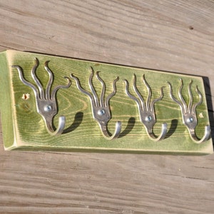 Könnte beinhalten: Ein handgefertigter Wand-Kleiderhaken. Der Haken besteht aus einer grün lackierten Holzplatte mit vier Metallgabeln, die als Haken angebracht sind. Die Gabeln sind mit Schrauben befestigt, und der Haken ist auf einer Holzoberfläche montiert.