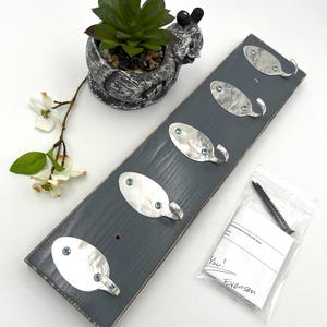 May include: A gray wooden wall-mounted coat rack with four silver spoon-shaped hooks. The hooks are attached with blue screws. A small bag of screws and a decorative succulent in a snail-shaped pot are also present.