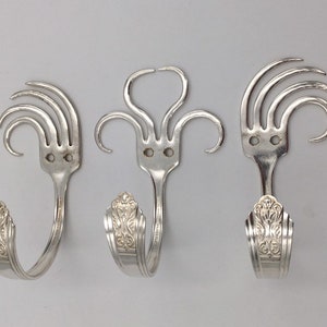 Può includere: Tre ganci a forma di forchetta argentati, ognuno con un design unico, sono montati su una parete bianca. Le forchette sono state piegate a forma di gancio, con i denti della forchetta che formano un motivo decorativo nella parte superiore.