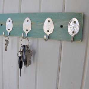May include: A teal blue wooden key rack with five hooks made from repurposed spoons. The hooks are attached to the wood with screws. There are keys hanging on three of the hooks.