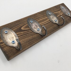 Könnte beinhalten: Ein Schlüsselhalter aus Holz mit vier Haken aus wiederverwendeten Löffeln. Die Haken sind mit Schrauben am Holz befestigt. Das Holz ist dunkelbraun.