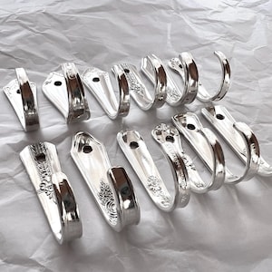 Op de afbeelding: Een verzameling van 13 zilverkleurige metalen haken gemaakt van hergebruikt bestek. De haken zijn allemaal in verschillende vormen en maten, en sommige hebben decoratieve patronen.