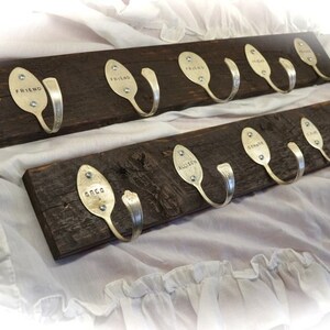 Puede incluir: Dos percheros rústicos de madera con ganchos de cuchara plateada. Cada perchero tiene múltiples ganchos hechos de cucharas reutilizadas. Algunas cucharas tienen nombres como "Friend" y "Anca" estampados. La madera es de color marrón oscuro.