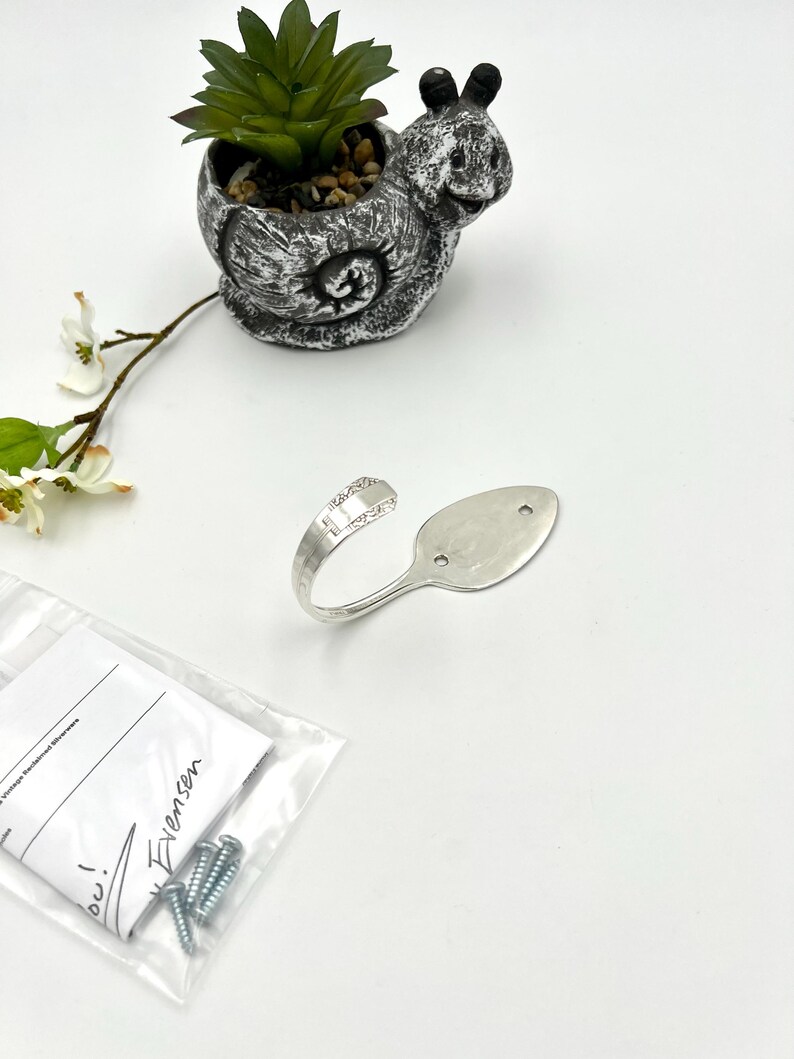 May include: A silver spoon repurposed into a decorative hook. The hook is next to a small snail-shaped planter holding a succulent plant. A small bag with screws and a handwritten note is also present.
