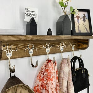 May include: A rustic wooden shelf with decorative fork hooks. The shelf holds a black framed picture, a small house-shaped vase, and a stack of books. Scarves and a purse hang from the fork hooks. The shelf is mounted on a white wall.