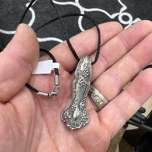 Puede incluir: Un collar con un colgante de cuchara de plata con un intrincado diseño floral, que cuelga de un cordón negro.