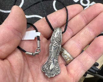 Puede incluir: Un collar con un colgante de cuchara de plata con un intrincado diseño floral, que cuelga de un cordón negro.