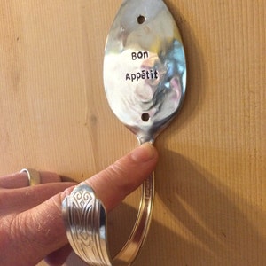 Peut inclure: Une cuill&egrave;re en argent pli&eacute;e en crochet avec les mots "Bon App&eacute;tit" grav&eacute;s sur la cuill&egrave;re. La cuill&egrave;re est mont&eacute;e sur un mur en bois.