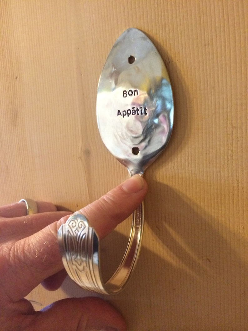 Peut inclure: Une cuill&egrave;re en argent pli&eacute;e en crochet avec les mots "Bon App&eacute;tit" grav&eacute;s sur la cuill&egrave;re. La cuill&egrave;re est mont&eacute;e sur un mur en bois.