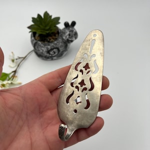 May include: A vintage silver cake server with a decorative openwork design. The server has a curved handle with an ornate pattern and a slotted blade. The server is held in a hand against a white background.