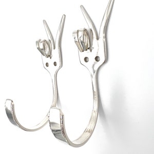 Peut inclure: Deux crochets en forme de U en argent, fabriqués à partir de fourchettes, sont fixés à un mur blanc. Les fourchettes sont placées côte à côte, les dents vers le haut.