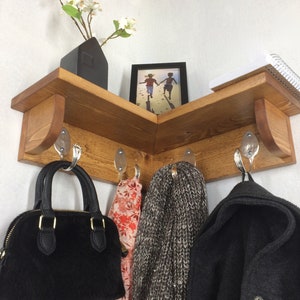 Pode incluir: Uma prateleira de canto de madeira com ganchos para pendurar objetos. A prateleira é tingida de uma cor marrom escura e tem um design simples. Há cinco ganchos na prateleira, e uma bolsa preta, um cachecol cinza e uma jaqueta preta estão pendurados neles. Um pequeno porta-retratos com uma foto de duas crianças está na prateleira, junto com um vaso de flores brancas.