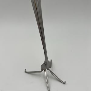 May include: A silver fork bent into a tripod stand. The fork is standing upright on a white surface.