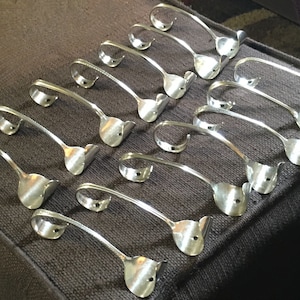 Puede incluir: Un conjunto de 15 ganchos de metal plateados hechos de cubiertos reutilizados. Los ganchos tienen forma de cuchara con el cuenco doblado en forma de gancho. Los ganchos están dispuestos en filas sobre una superficie de tela gris.