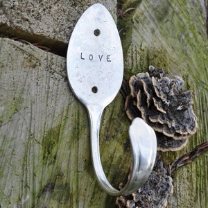 Peut inclure: Une cuill&egrave;re en argent transform&eacute;e en crochet avec le mot "LOVE" grav&eacute; sur le devant. Le crochet est mont&eacute; sur une surface en bois avec une croissance en forme de champignon.
