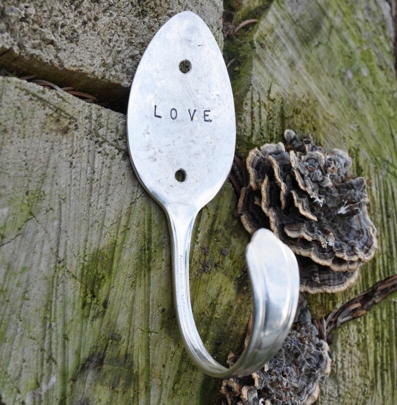 Peut inclure: Une cuill&egrave;re en argent transform&eacute;e en crochet avec le mot "LOVE" grav&eacute; sur le devant. Le crochet est mont&eacute; sur une surface en bois avec une croissance en forme de champignon.