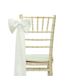May include: A cream-colored Chiavari chair with a white satin bow tied to the back. The chair has a padded seat and a bamboo-style frame, ideal for weddings and events.