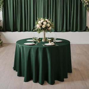 Könnte beinhalten: Eine runde, dunkelgrüne Tischdecke ist auf einem Tisch mit weißen Tellern, Besteck und Gläsern gedeckt. Eine Vase mit weißen Blumen steht in der Mitte des Tisches.