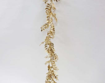 May include: A gold-colored, artificial garland with leaf-shaped ornaments. The garland is hanging vertically against a white background. The leaves are detailed and have a metallic sheen, suggesting a decorative or festive purpose.