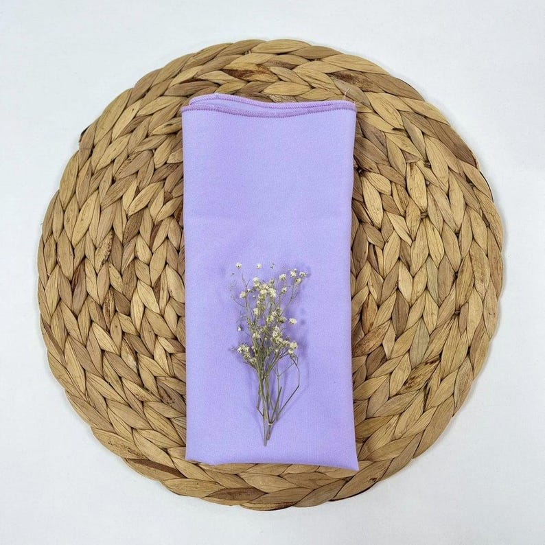 May include: A lavender-coloured cloth napkin on a woven straw placemat with a small bouquet of white flowers.