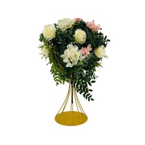 May include: A floral arrangement featuring white roses, pink and white flowers, and green foliage. The arrangement is supported by a gold-colored metal stand with a circular base and a series of vertical supports.