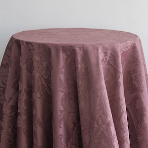 May include: A round, mauve tablecloth featuring a detailed floral pattern. The fabric drapes over a table, displaying the intricate design and texture. The tablecloth is made of a soft, luxurious material.