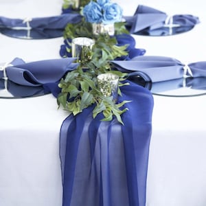 May include: A blue chiffon table runner with greenery and silver candle holders. The runner is draped over a white tablecloth and there are blue napkins on the table.