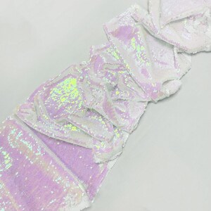 Op de afbeelding: Een stuk iriserende stof met witte en lavendelkleurige pailletten. De stof is gevouwen en gedrapeerd, wat het glinsterende effect van de pailletten laat zien. De pailletten reflecteren een reeks kleuren, waaronder groen en geel.