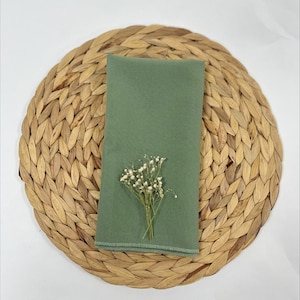 May include: A sage green linen napkin with a small bouquet of white flowers on top, placed on a woven straw placemat.