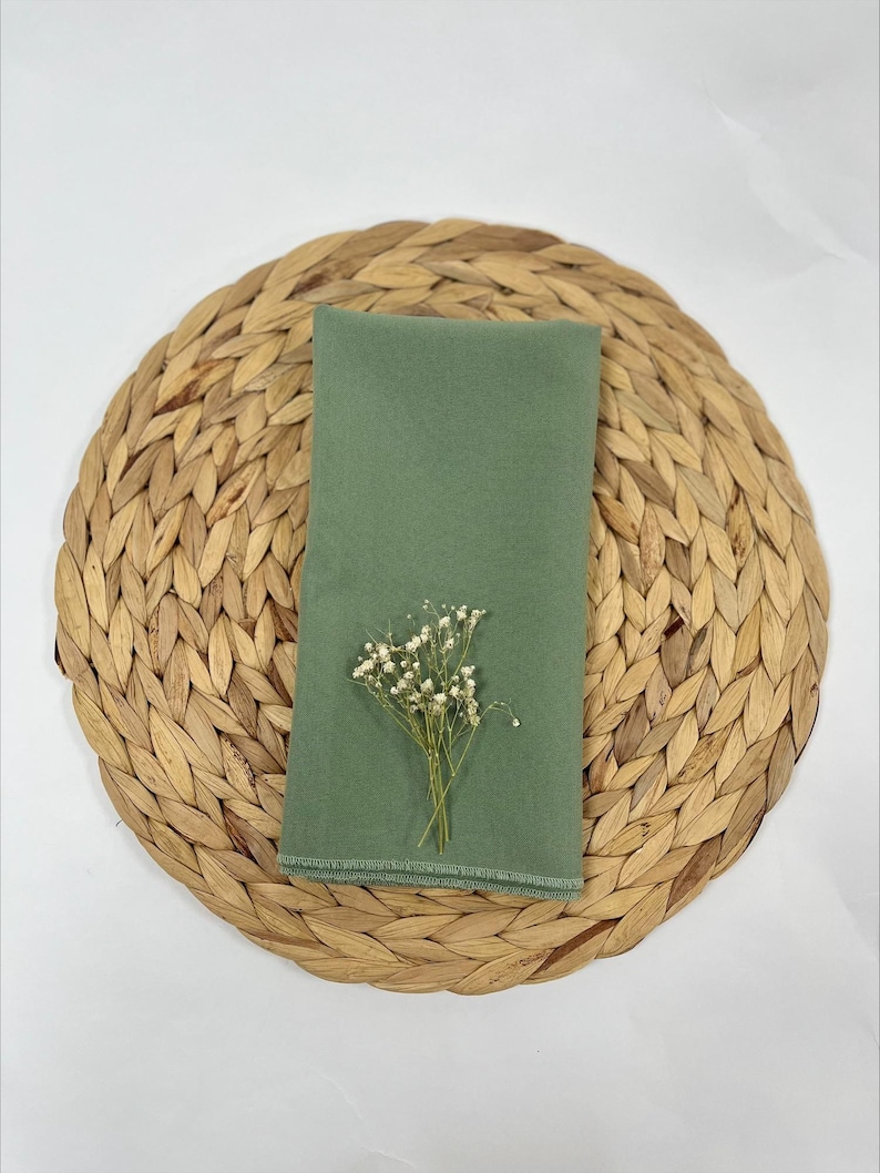 May include: A sage green linen napkin with a small bouquet of white flowers on top, placed on a woven straw placemat.