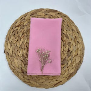 May include: A pink fabric napkin on a woven round placemat with a small bouquet of white flowers.