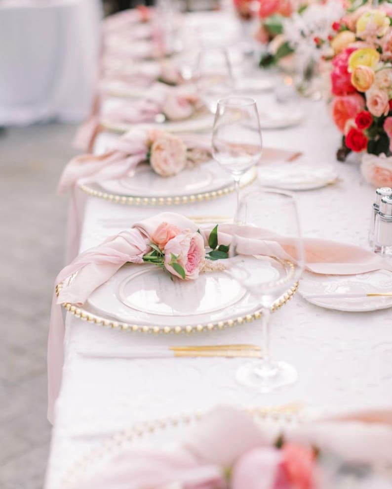 May include: A white table setting with a pink napkin and a single pink rose on a white plate with a gold beaded rim.