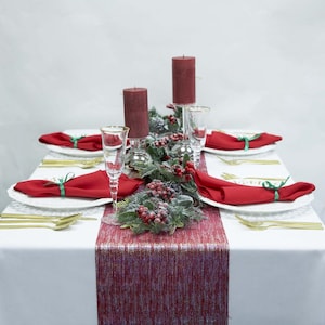 May include: A festive table setting with a white tablecloth, featuring red napkins, gold cutlery, and white plates. The centerpiece includes burgundy candles, greenery, and a red table runner. Champagne flutes and decorative accents complete the holiday theme.