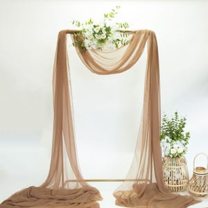 May include: A brown sheer fabric backdrop for a wedding ceremony or event. The fabric is draped over a metal archway and decorated with white flowers.