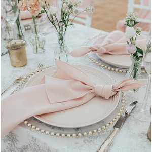 May include: A close-up of a wedding table setting with a pink napkin folded into a bow on a white plate with a gold beaded charger. The table is covered in a white tablecloth with a grey floral pattern. There are two small vases with pink and white flowers.