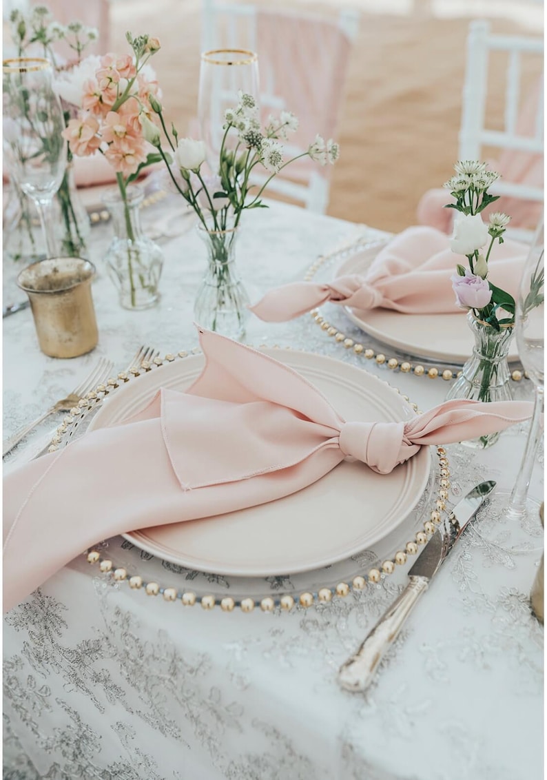 May include: A close-up of a wedding table setting with a pink napkin folded into a bow on a white plate with a gold beaded charger. The table is covered in a white tablecloth with a grey floral pattern. There are two small vases with pink and white flowers.
