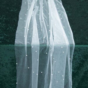 May include: White tulle fabric with small white pearl embellishments. The fabric is draped over a green surface.