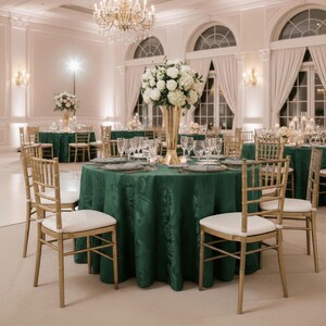 May include: Elegant banquet hall with round tables set for a formal event. Each table is covered with a dark green tablecloth and adorned with white floral centerpieces in gold vases. Gold chairs with white cushions surround the tables.