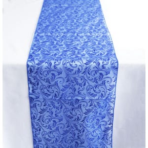 May include: A vibrant blue table runner with a repeating floral design. The runner, made of a shiny material, is laid on a white tablecloth. The pattern features swirling leaves and vines, adding elegance.