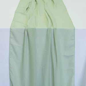 May include: A pale green table runner made from a soft, flowing material. The runner is draped over a white surface, creating soft folds and shadows. The fabric has a subtle sheen, adding a touch of elegance. Suitable for weddings or special occasions.
