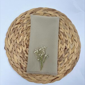 May include: A light beige linen napkin with a small bouquet of white flowers on top of it. The napkin is sitting on a woven straw placemat.