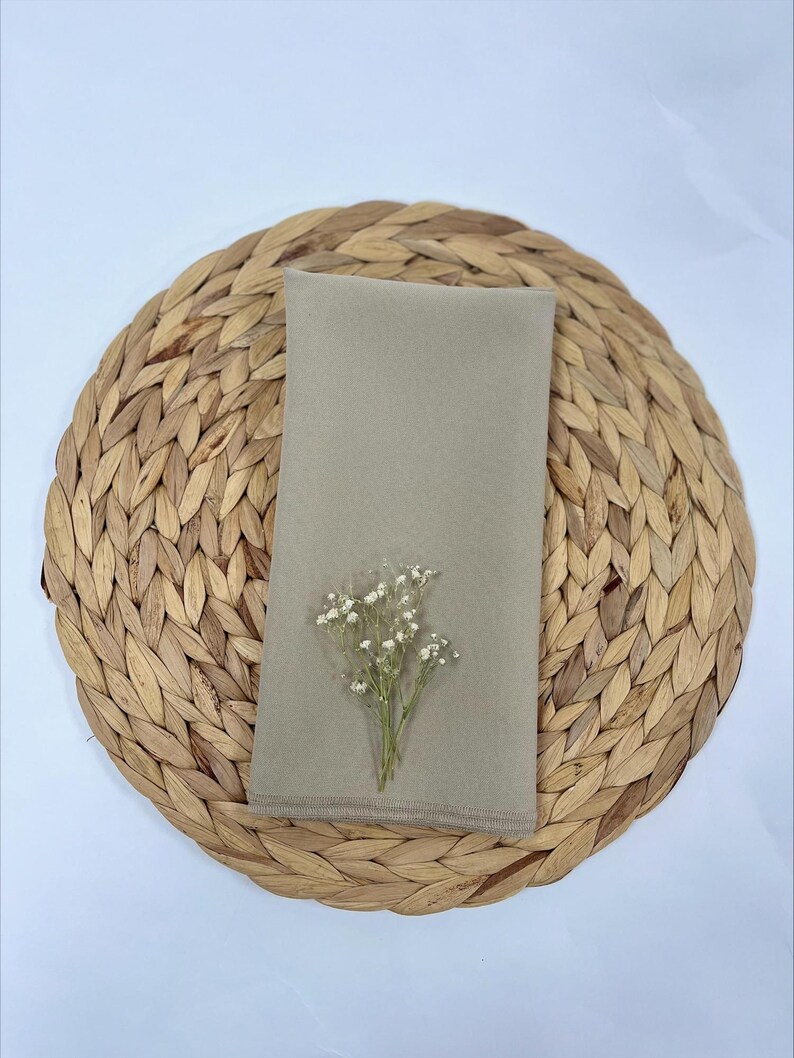 May include: A light beige linen napkin with a small bouquet of white flowers on top of it. The napkin is sitting on a woven straw placemat.