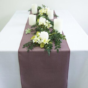 May include: A table setting with a plum-coloured table runner, white pillar candles, and floral arrangements. The flowers include white roses and yellow blossoms, with green foliage. The table is covered with a white tablecloth.