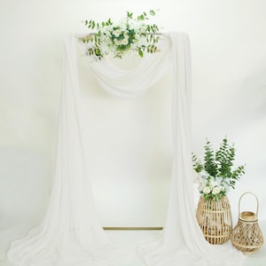 May include: A white wedding arch with draped fabric and floral arrangements. The arch is decorated with white flowers and greenery. A woven basket with flowers and a decorative lantern are on the side.