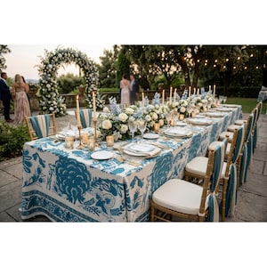 Puede incluir: Una mesa larga preparada para un evento formal al aire libre. La mesa está cubierta con un mantel estampado en azul y blanco y está decorada con platos con borde dorado, cubiertos y cristalería. Los centros de mesa florales y las velas adornan la mesa.