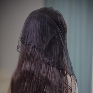 May include: A black veil with a scalloped lace edge is draped over long, dark brown hair. The veil is sheer, allowing the hair to be partially visible. The background is a soft gray and blue.